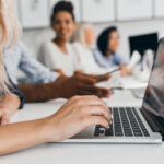 Blonde woman with elegant hairstyle typing text on keyboard in office. Indoor portrait of international employees with secretary using laptop on foreground..